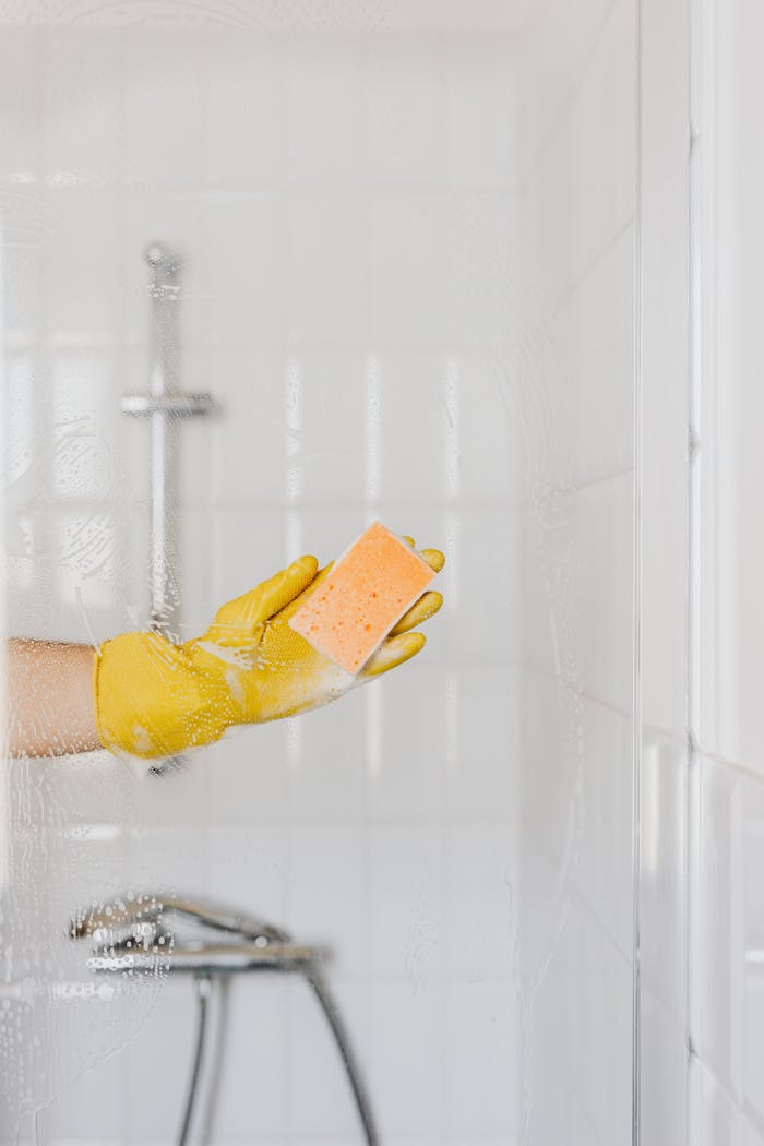 A close-up of a gloved hand holding a sponge cleaning a glass shower door.